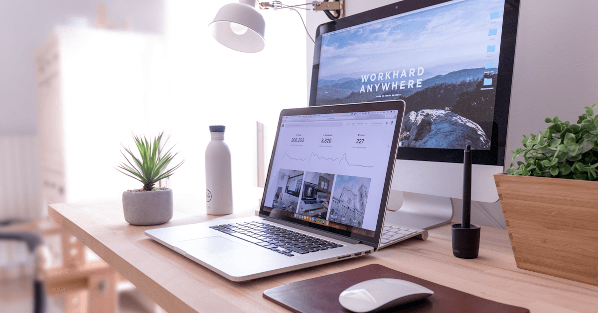 Scrivania da lavoro con laptop aperto e monitor acceso, bottiglia d'acqua e pianta, luce naturale del mattino