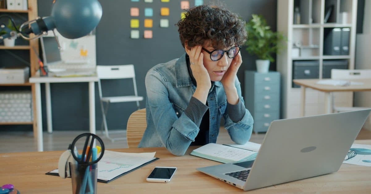 Professionista con occhiali si massaggia le tempie davanti al laptop in un ufficio luminoso, segno di stanchezza e pressione lavorativa
