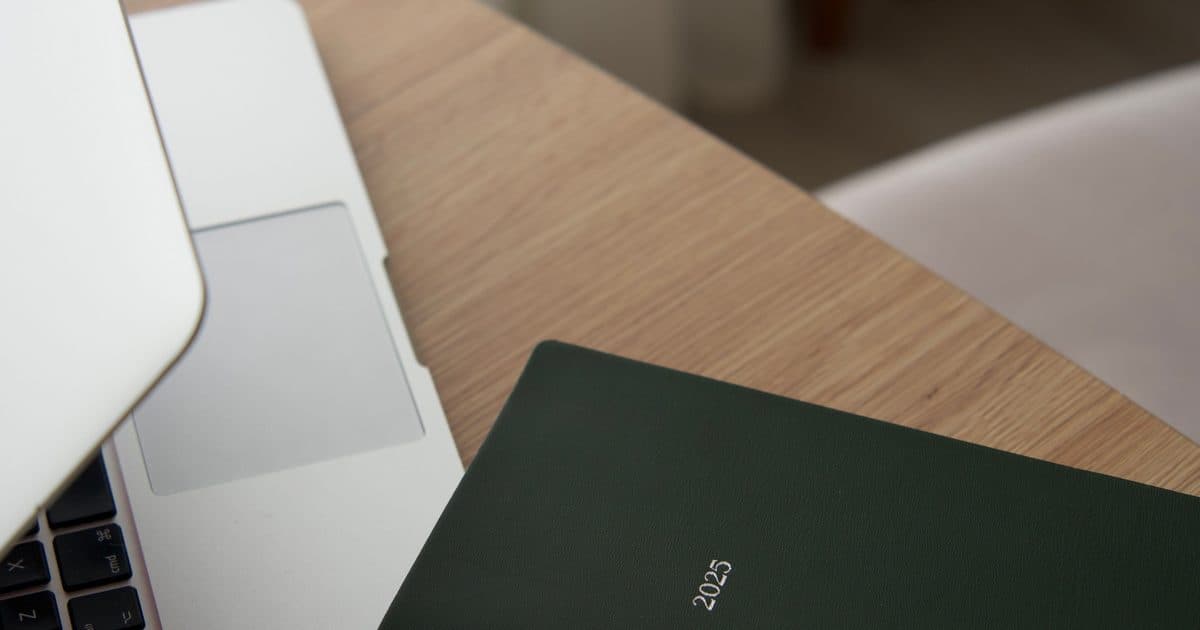 Scrivania in legno chiaro con un laptop e un'agenda 2025 dalla copertina verde scuro, luce naturale del mattino, a rappresentare un piano scritto in anticipo per le settimane a bassa energia