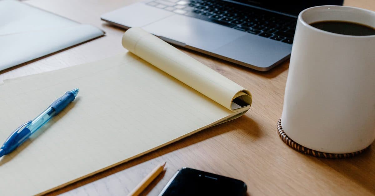 Taccuino aperto, penna e tazza di caffè su una scrivania in legno accanto a un laptop, spazio di lavoro ordinato